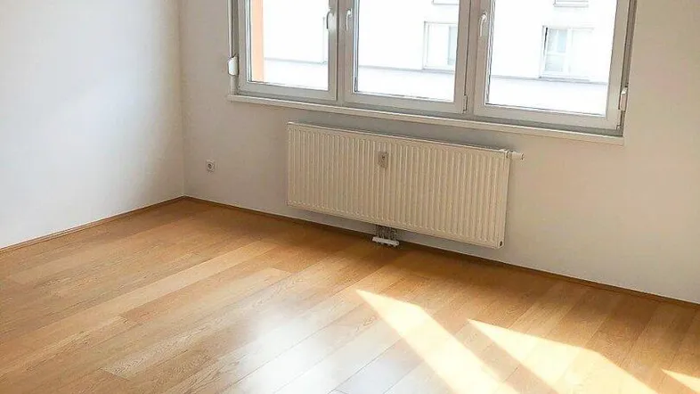 Helles Zimmer mit Holzparkettboden, weißen Wänden und großem Fenster, das viel Tageslicht hereinlässt.