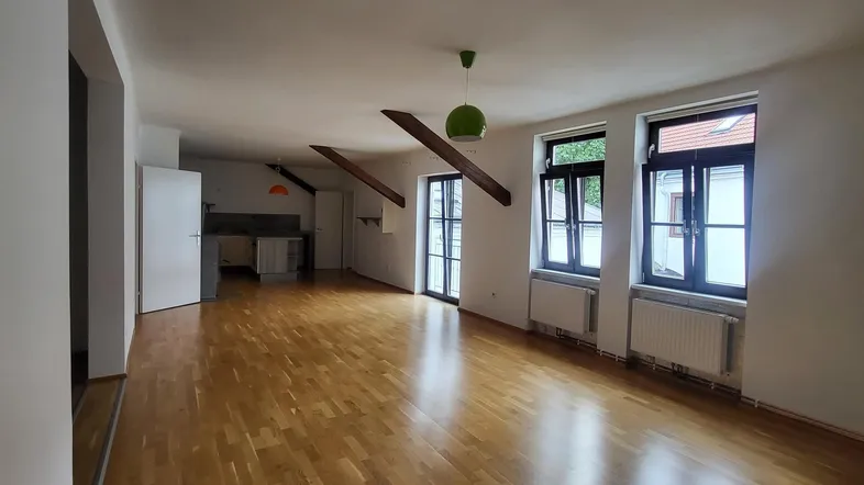 Geräumiger Wohnbereich mit hellem Parkettboden, sichtbaren Holzbalken und großen Fenstern, die viel Tageslicht hereinlassen.