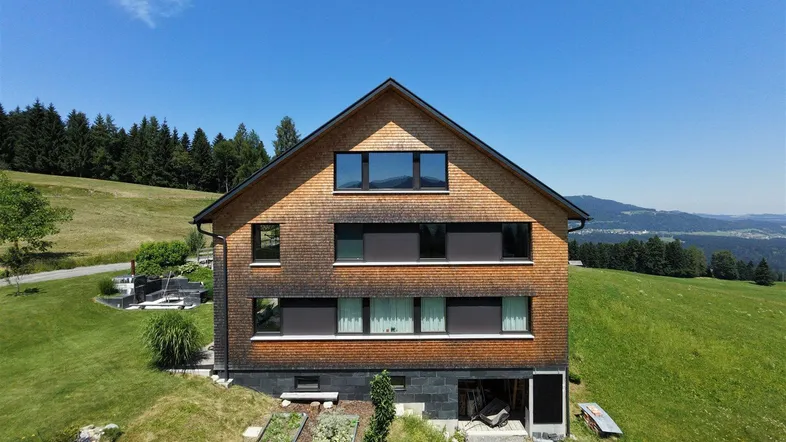 Modernes Haus mit Holzfassade und Satteldach in ländlicher Umgebung mit Bergblick und Gartenbeeten.