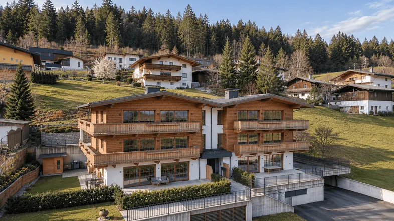 Modernes Chalet mit Holzfassade, mehreren Balkonen und Terrasse in alpiner Hanglage mit Waldblick.