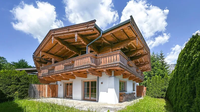 Traditionelles Chalet mit charakteristischen Holzbalkonen, weißer Fassade und gepflegtem Gartenbereich unter blauem Himmel.
