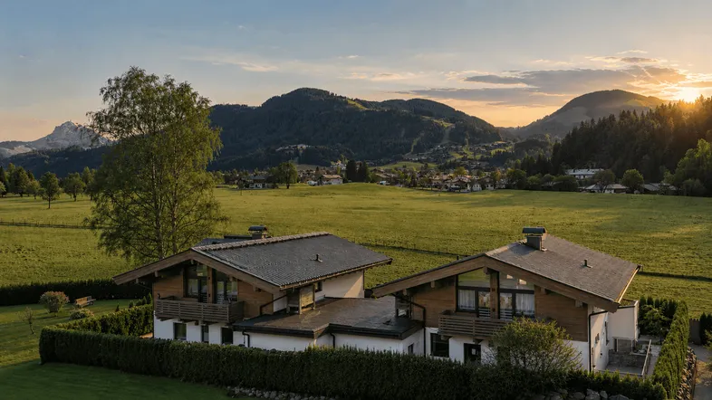 Panoramablick auf zwei moderne Chalets in einer grünen Wiesenlandschaft mit Bergen und Sonnenuntergang.