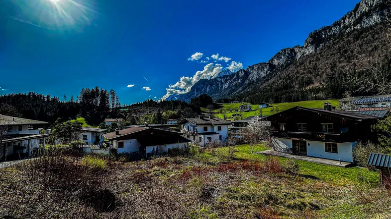 Sonnige Alpenlandschaft mit traditionellen Häusern, grünen Wiesen und majestätischen Bergen im Hintergrund.