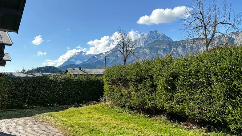 Grüner Gartenbereich mit gepflastertem Weg, Hecke und beeindruckendem Bergblick unter blauem Himmel.