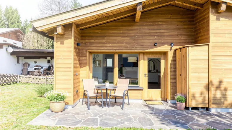 Gemütliche Steinterrasse mit Sitzgelegenheiten vor einem Holzhaus, umgeben von Grün.