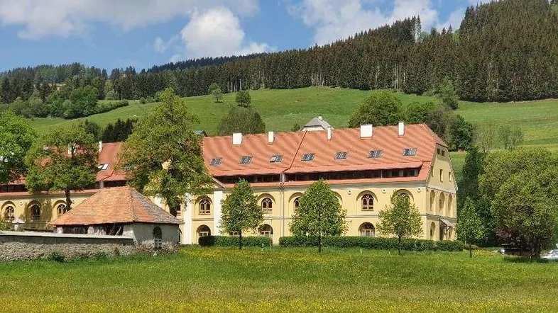 Historisches Gebäude mit gelber Fassade und rotem Dach, umgeben von grünen Wiesen und Bäumen.