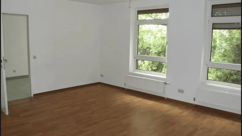 Helles Zimmer mit Parkettboden und zwei großen Fenstern, die einen Blick ins Grüne bieten.