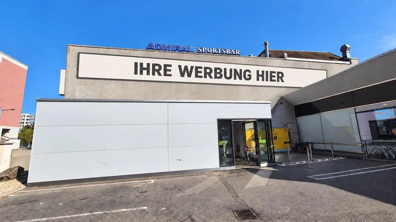 Modernes Geschäftsgebäude mit großer Werbefläche und Eingangsbereich, umgeben von einem Parkplatz.