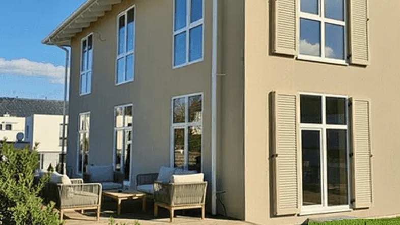Moderne Hausfassade mit beiger Putzfassade, großen Fenstern und einer Holzterrasse mit Sitzgelegenheiten.