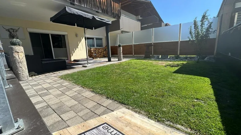 Sonniger Außenbereich mit gepflasterter Terrasse, Rasenfläche und Holzlager.