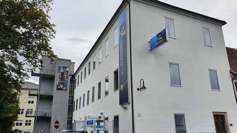 Weißes, gut erhaltenes Gebäude mit mehreren Fenstern und Werbebannern an der Fassade, umgeben von parkenden Autos.