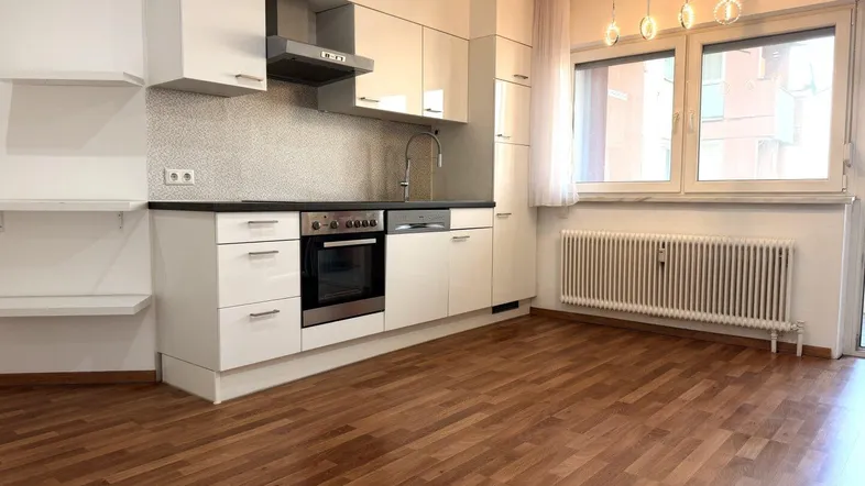 Moderne Einbauküche mit weißen Hochglanzfronten, dunkler Arbeitsplatte und Holzoptikboden in einem hellen Raum.