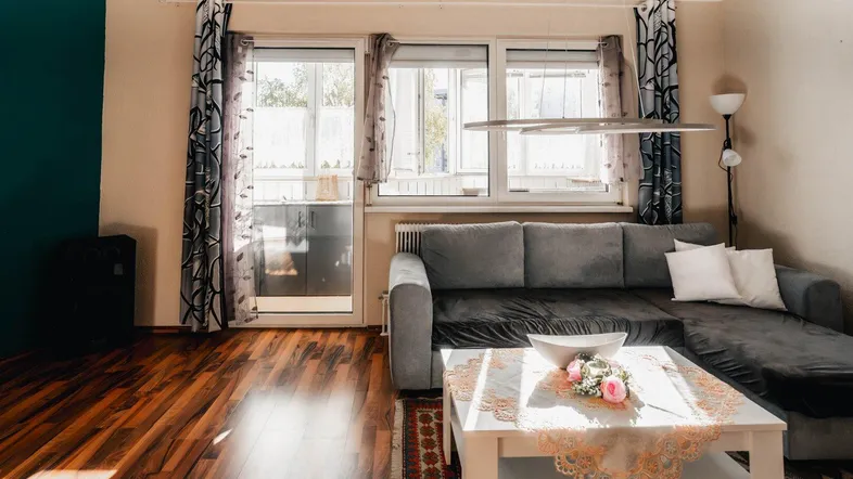 Helles Wohnzimmer mit grauem Ecksofa, Holzboden und Zugang zum Balkon, gemütlich eingerichtet.