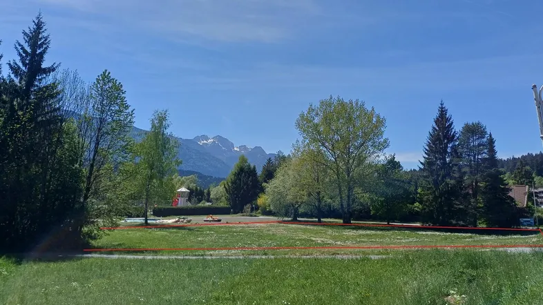 Weitläufige Grünfläche mit Bergblick, umgeben von Bäumen und einem kleinen Spielplatz.