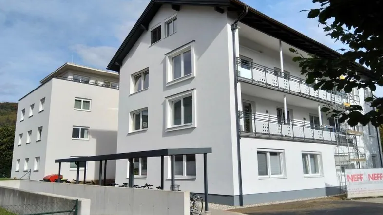 Weißes Mehrfamilienhaus mit Balkonen und überdachtem Eingangsbereich unter blauem Himmel.