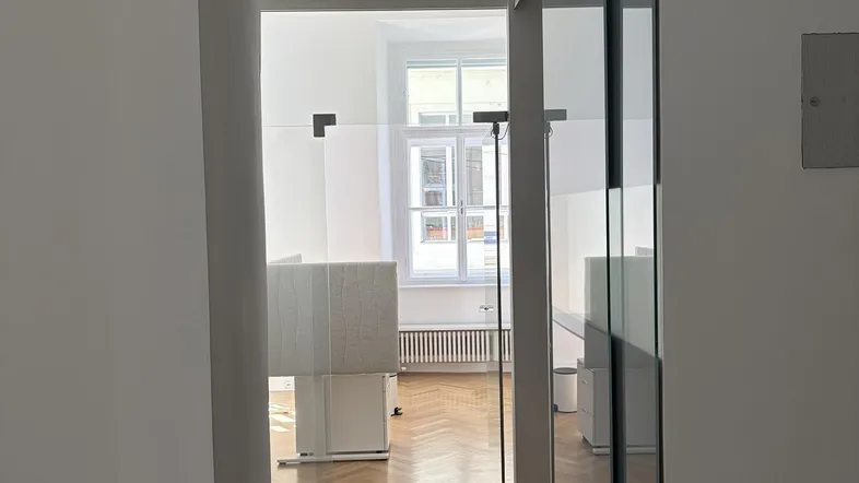 Blick durch eine moderne Glastür in ein helles Büro mit Fischgrätparkett und großen Fenstern.