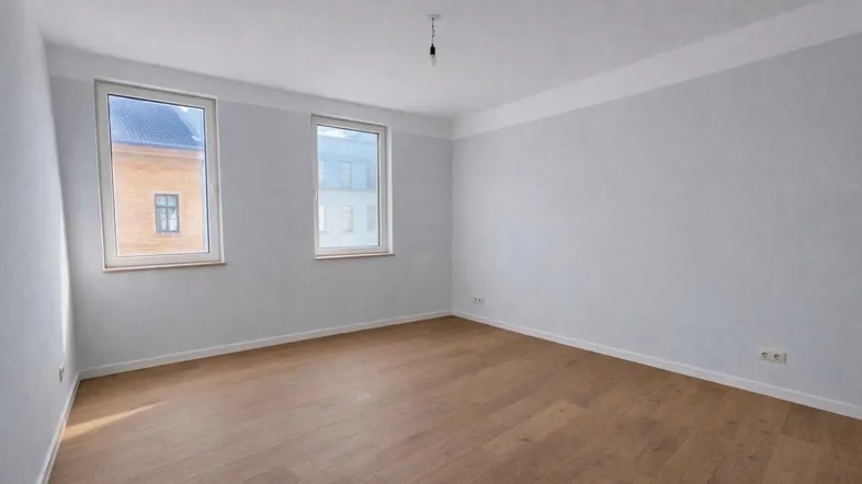 Leerer Raum mit zwei Fenstern, Holzboden und weißen Wänden, ideal für Schlaf- oder Wohnbereich.