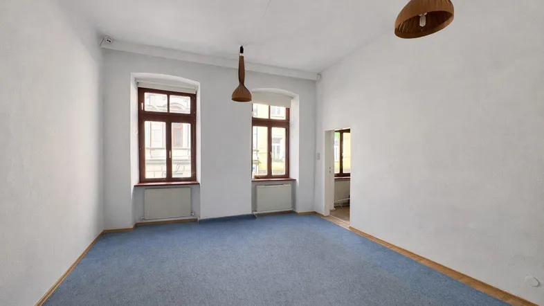 Heller, leerer Wohnraum mit blauem Teppichboden, hohen Decken und zwei Fenstern, die Tageslicht hereinlassen.