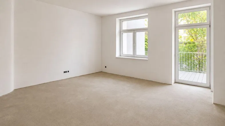 Helles, unmöbliertes Zimmer mit weiß gestrichenen Wänden, Betonboden und Zugang zu einem kleinen Balkon.
