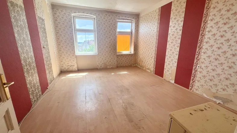 Helles Wohnzimmer in einem Altbau mit abgenutztem Dielenboden und floraler Tapete, das eine Renovierung benötigt.