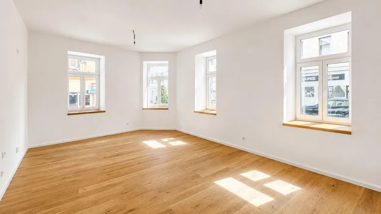 Helles, leeres Zimmer mit Holzboden und mehreren Fenstern, durch die Sonnenlicht einfällt.