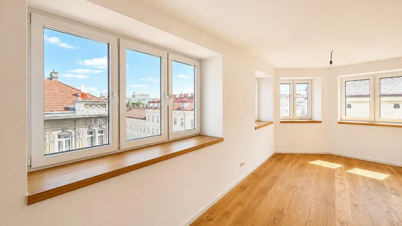 Helles, leeres Zimmer mit Holzboden und mehreren Fenstern, die einen Blick auf die Stadt bieten.