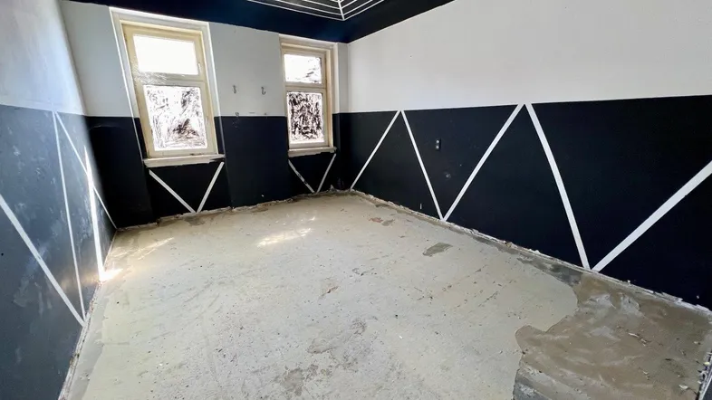 Leeres Zimmer in Renovierung mit auffälliger schwarz-weißer geometrischer Wand- und Deckengestaltung und zwei Fenstern.