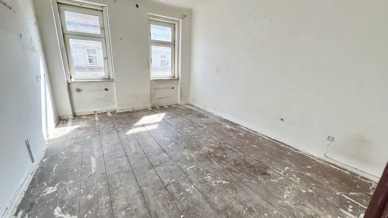 Leeres Zimmer mit abgenutztem Dielenboden und zwei Fenstern, bereit zur Renovierung.