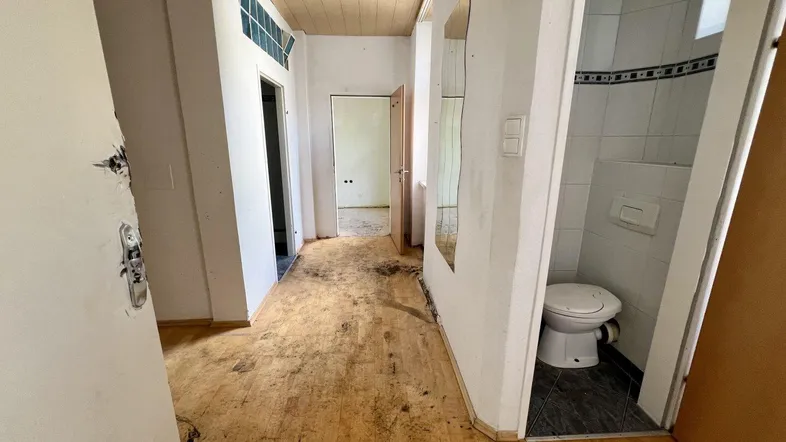 Langer Flur mit abgenutztem Holzboden und Blick auf eine separate Toilette, die renovierungsbedürftig ist.