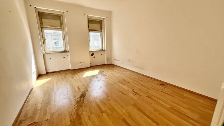 Leeres Zimmer mit Holzboden und zwei Fenstern, das Renovierungsbedarf aufweist.