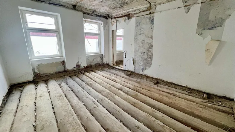 Unfertiger Raum mit freigelegten Holzdielen, zwei Fenstern und sichtbaren Deckenstrukturen, wartet auf Ausbau.