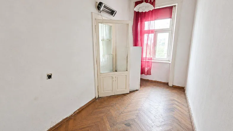 Leeres Zimmer mit Fischgrätparkett, weißen Wänden und rotem Vorhang am Fenster.