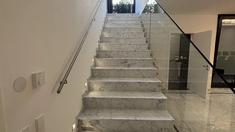 Moderne Treppe mit Marmorstufen, Glasgeländer und integrierter Beleuchtung, die zum Eingang führt.