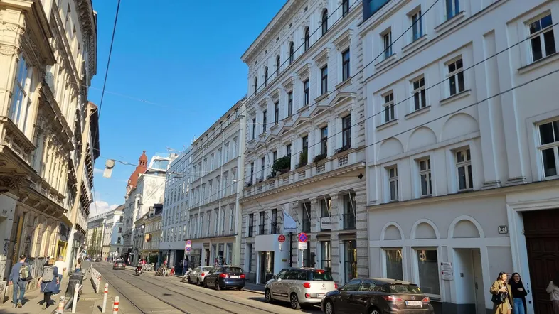 Historische Gebäude entlang einer belebten Straße in der Stadt, mit geparkten Autos und Fußgängern.