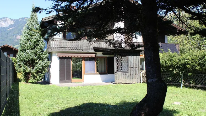 Ansicht des Hauses von hinten mit Garten, Terrasse und Balkon, umgeben von Bäumen und Bergen.