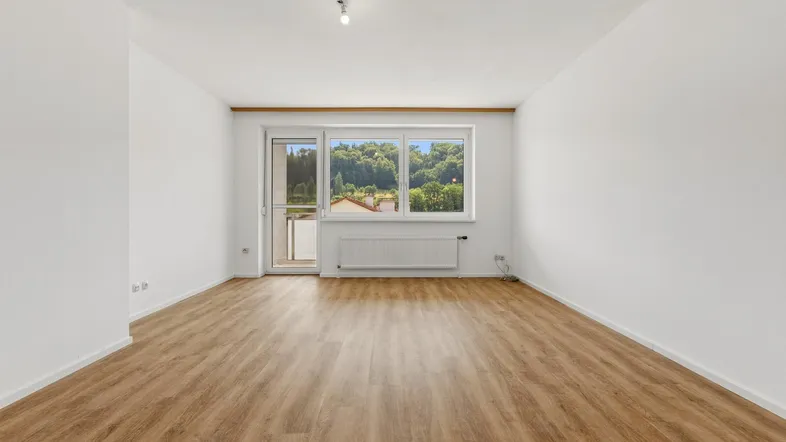 Helles Wohnzimmer mit Laminatboden, weißen Wänden und Zugang zu einem Balkon mit grüner Aussicht.