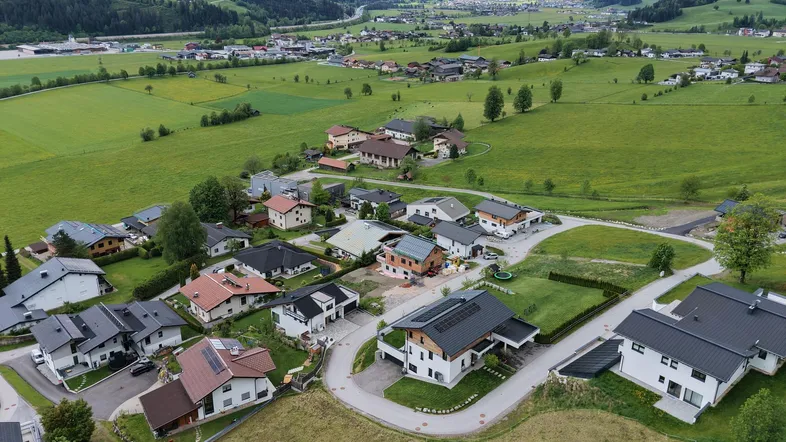 Luftaufnahme eines Wohngebiets in einem grünen Tal, umgeben von Bergen und Wiesen.