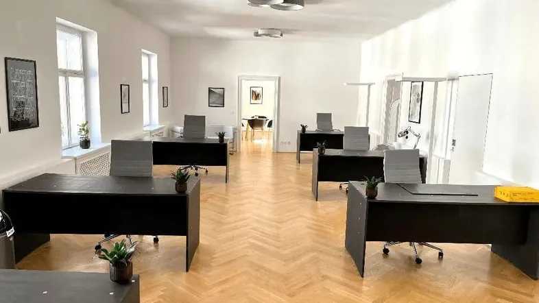 Geräumiges Büro mit mehreren Arbeitsplätzen, Fischgrätparkett und großen Fenstern, die viel Tageslicht hereinlassen.