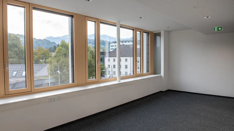 Heller Büroraum mit großen Holzfenstern, grauem Teppichboden und Blick auf die umliegende Landschaft.