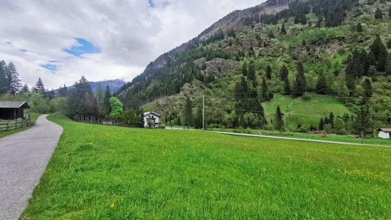 Weitläufige grüne Wiese mit einem Schotterweg und Blick auf bewaldete Berge unter bewölktem Himmel.