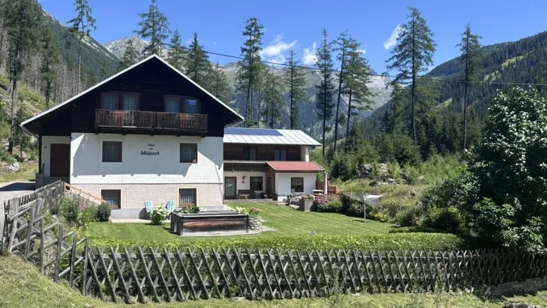 Traditionelles Chalet mit weißer Fassade, Holzbalkon und gepflegtem Garten, umgeben von einer beeindruckenden Berglandschaft.