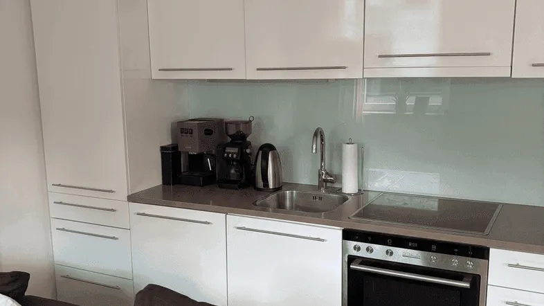 Moderne Küchenzeile mit weißen Hochglanzschränken, integriertem Backofen und Kaffeemaschine, angrenzend an den Wohnbereich.