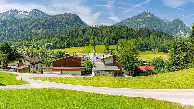 Panoramablick auf ein idyllisches Dorf mit Kirche, umgeben von grünen Bergen und Wäldern unter blauem Himmel.