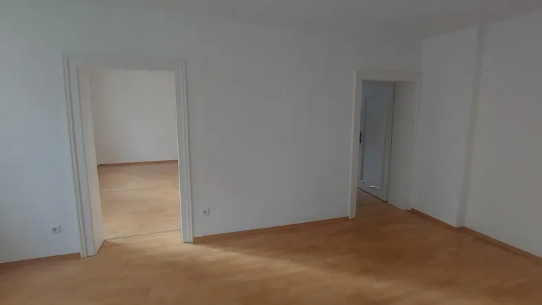 Leerer Raum mit hellem Holzfußboden und weißen Wänden, der Zugänge zu weiteren Zimmern bietet.