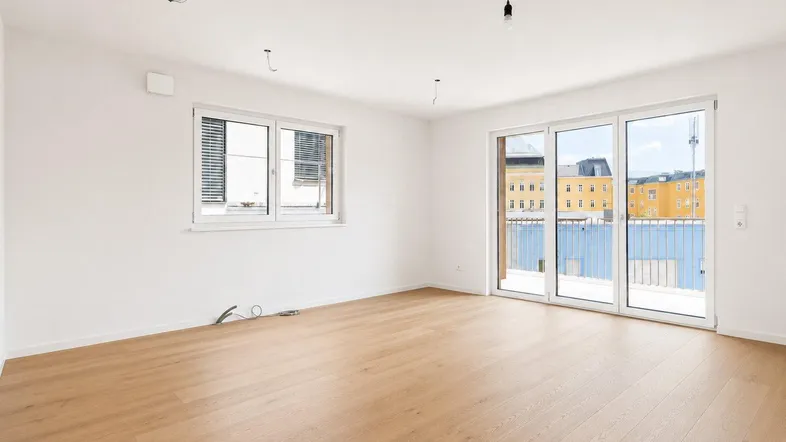 Heller, leerer Wohnraum mit Holzboden, großen Fenstern und Zugang zu einem Balkon mit Stadtblick.
