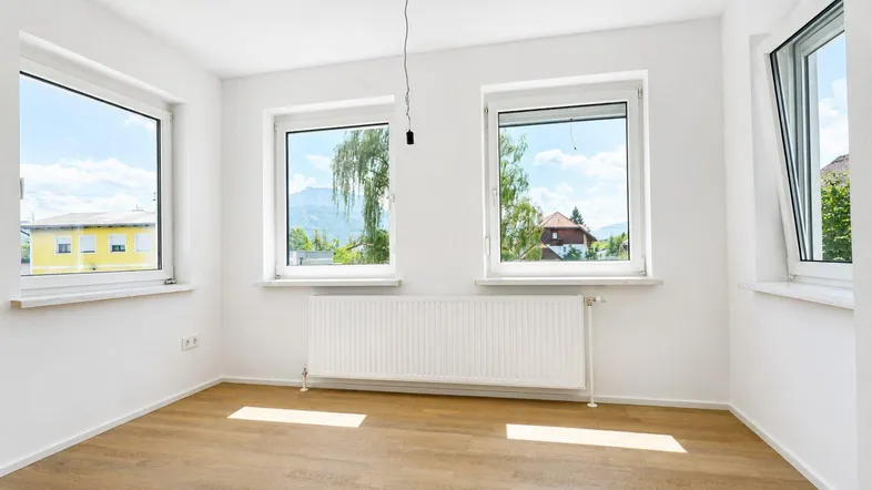 Helles Zimmer mit Parkettboden, weißen Wänden und Blick auf die Berge und Bäume.