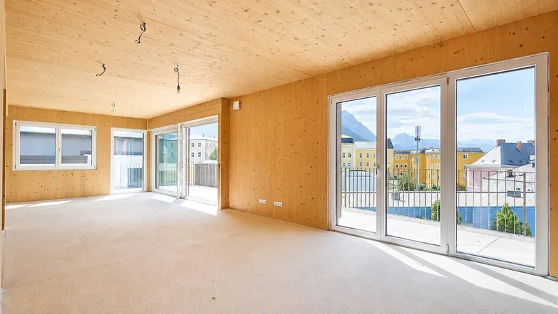 Heller, unfertiger Wohnraum mit Holzwänden, Holzbalkendecke und großen Fenstern mit Stadt- und Bergblick.