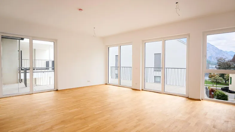 Helles, leeres Wohnzimmer mit Parkettboden, großen Fenstern und Bergblick.