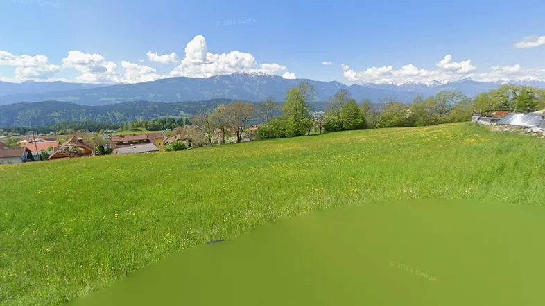 Weitläufige grüne Wiese mit Blick auf die umliegende Berglandschaft und eine Siedlung.