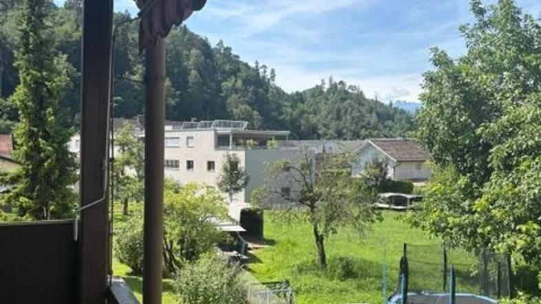 Blick vom Balkon auf den grünen Garten, umliegende Häuser und bewaldete Hügel unter blauem Himmel.